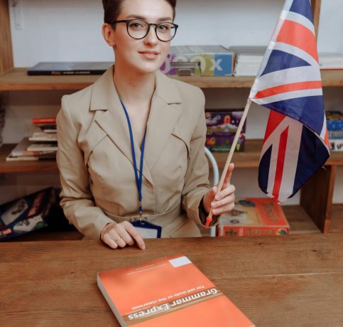 Portrait of a woman with eyeglasses holding a Union Jack flag at a desk.